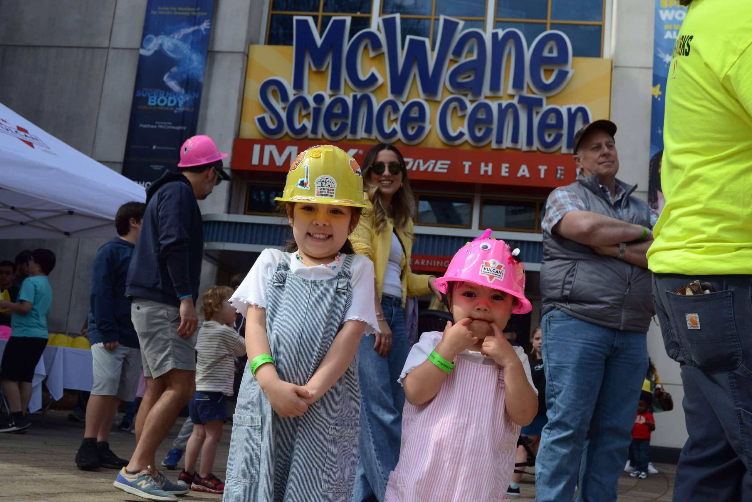 Two children wearing colorful construction hard hats smile outside McWane Science Center during Big Machines Day, surrounded by families