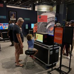 Child uses touchscreen at interactive AI exhibit in children's science center while families observe and learn.