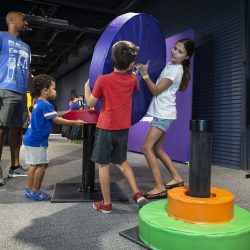 Kids and an adult explore a hands-on science center exhibit, moving giant foam rings in a Towers of Hanoi puzzle.