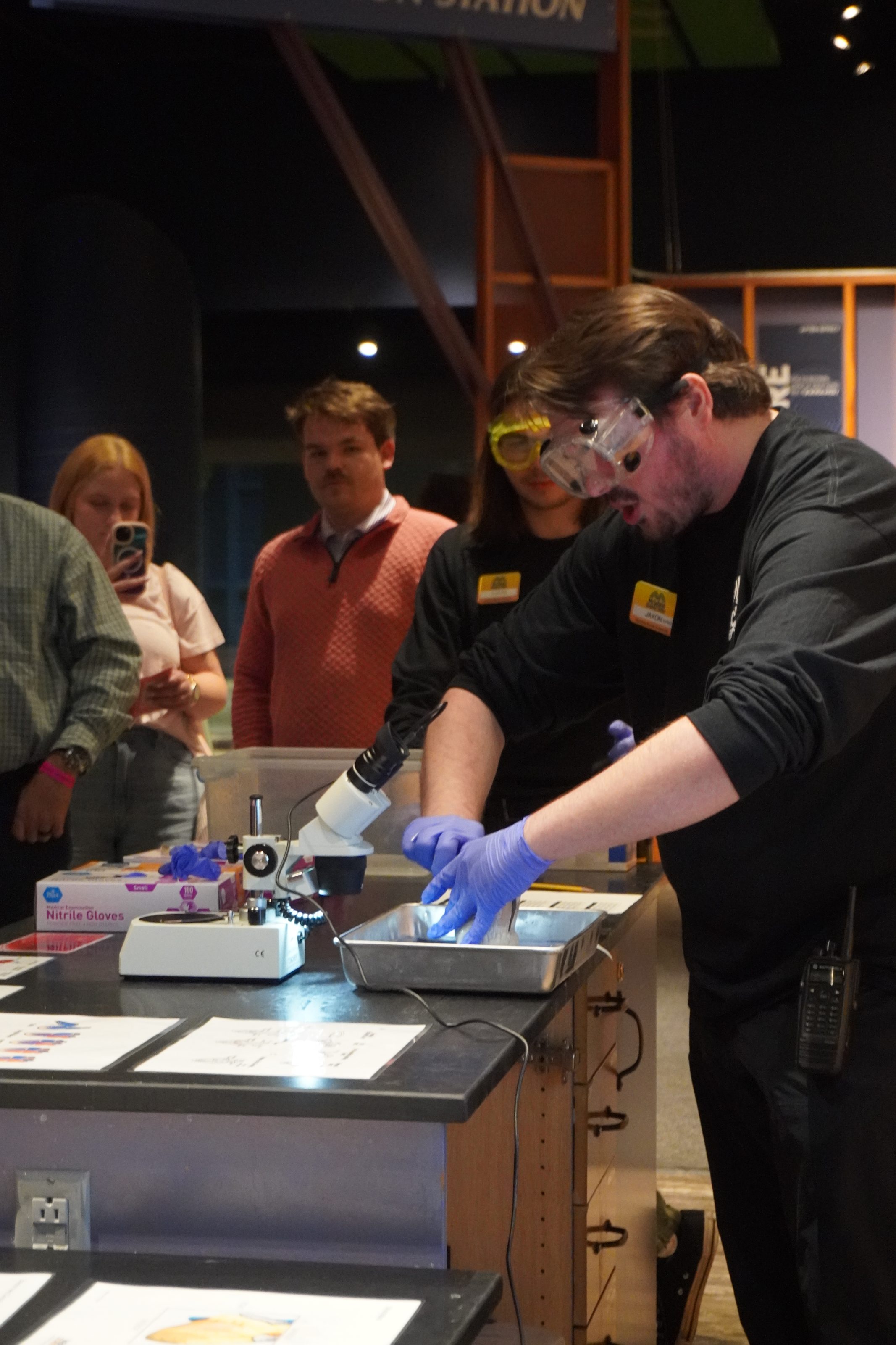An educator in safety goggles demonstrates a hands-on lab with microscope at a children's science center, visitors watching