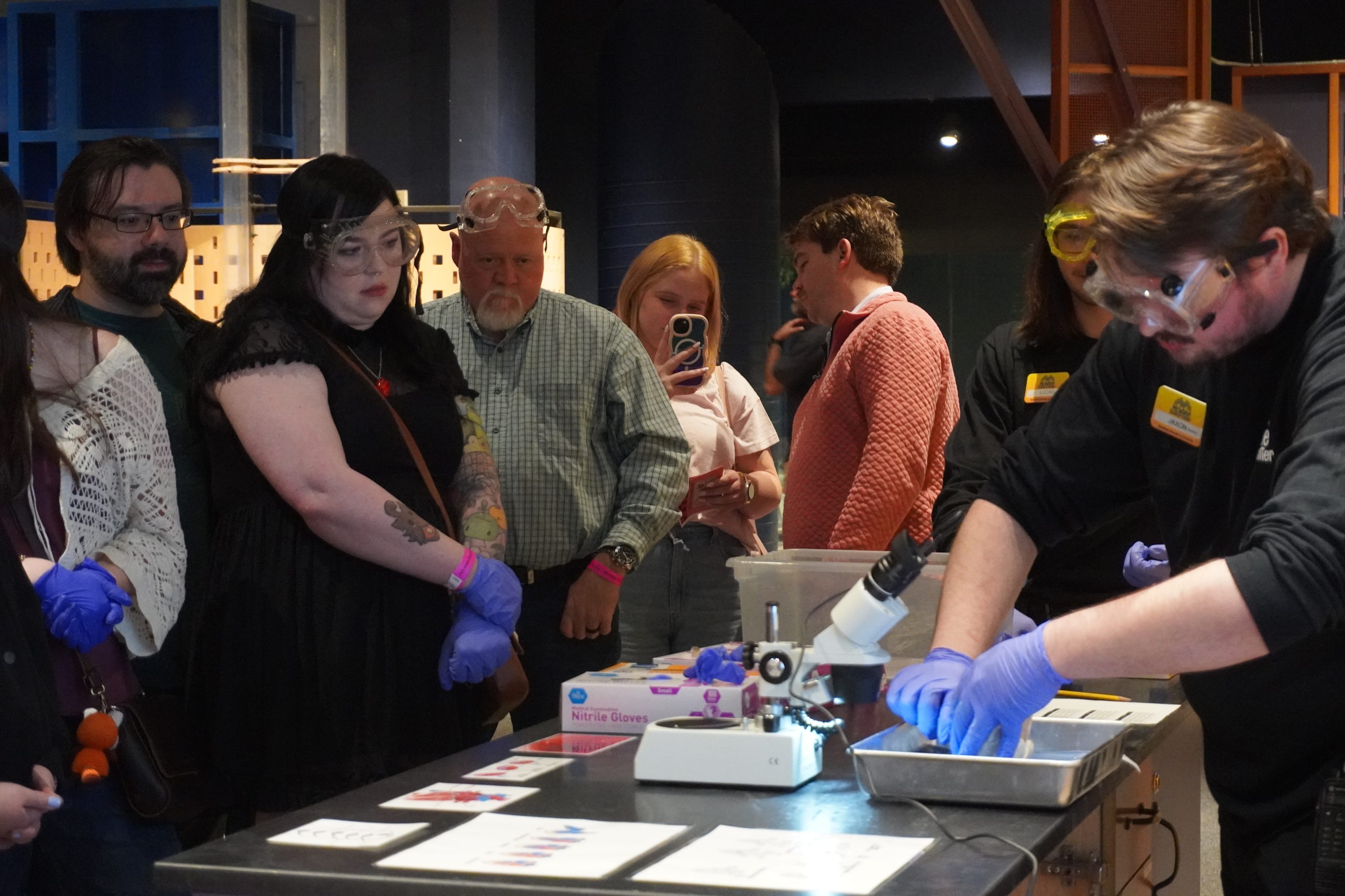 Visitors watch a science educator with gloves demonstrate a microscope experiment at a children's science center STEM lab.