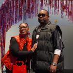 A woman and a man smile under a red tinsel arch at a children's science center event.