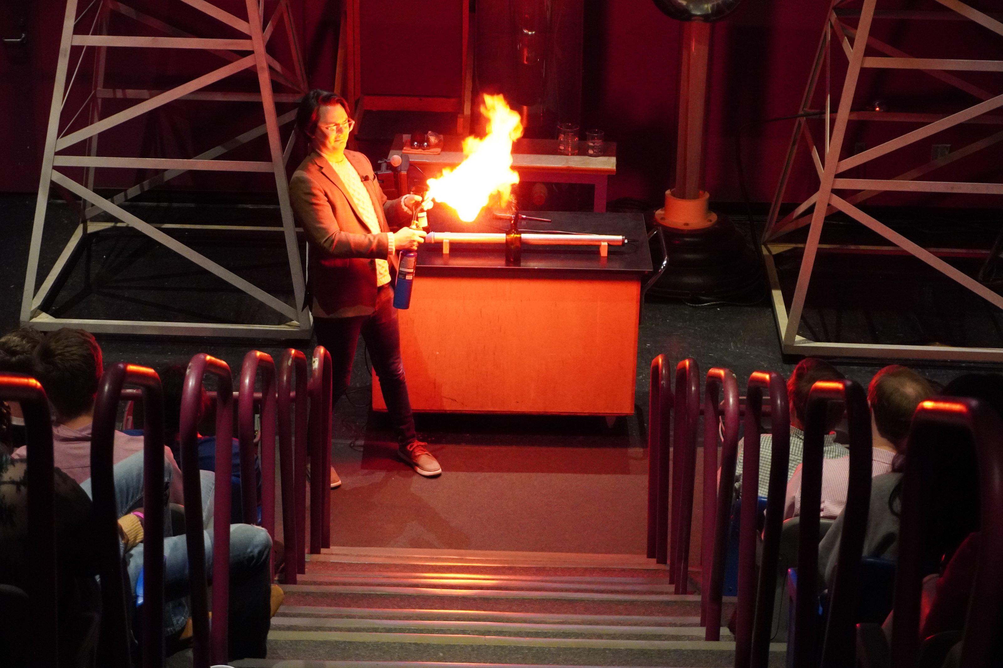 An educator ignites a fire experiment on stage during a children's science center show, thrilling the seated audience.