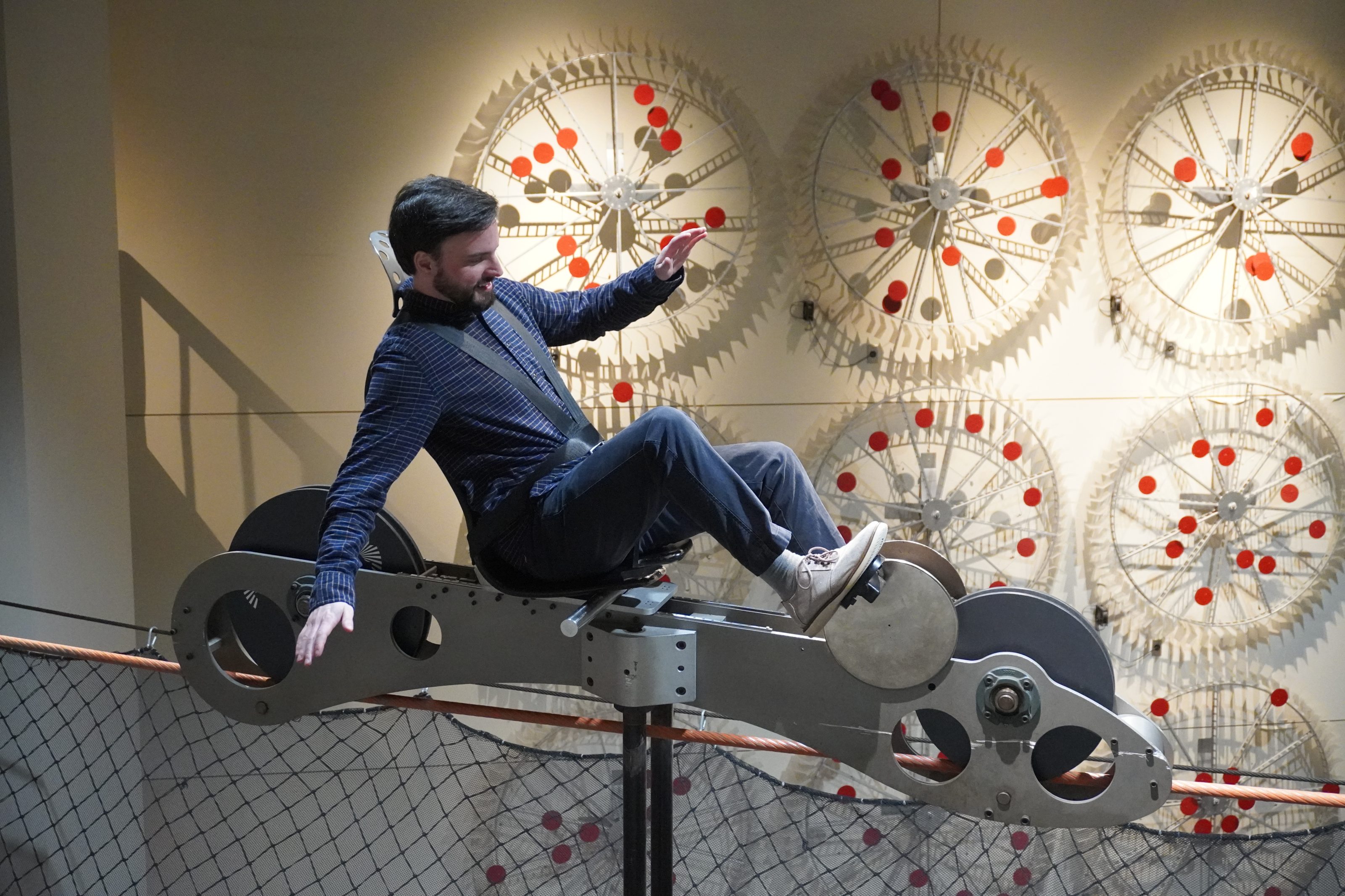A person tests a hands-on physics balance ride at a children's science center, with giant gear wheels in the background.