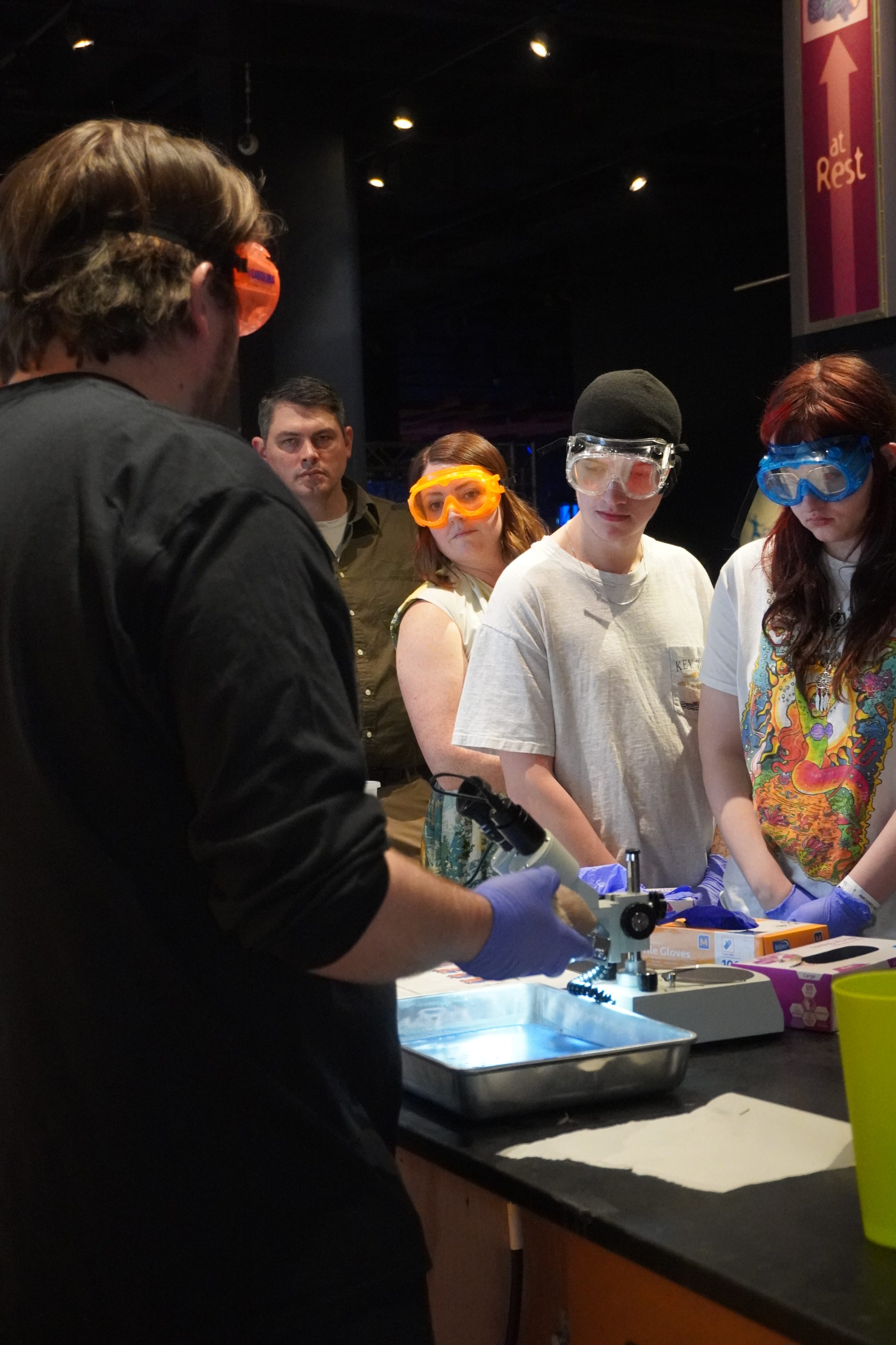 Students in safety goggles at children's science center watch a hands-on microscope experiment demo