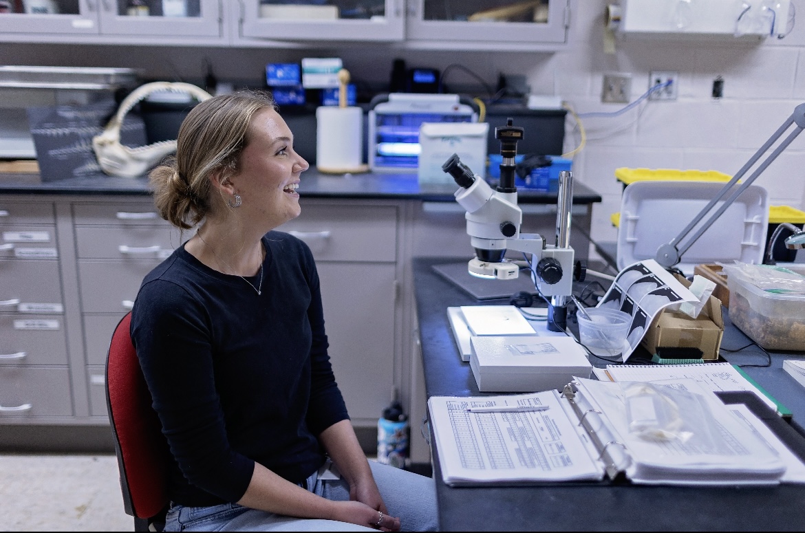 A woman scientist smiles beside a microscope in a STEM lab at a children's science center, with notes and samples.