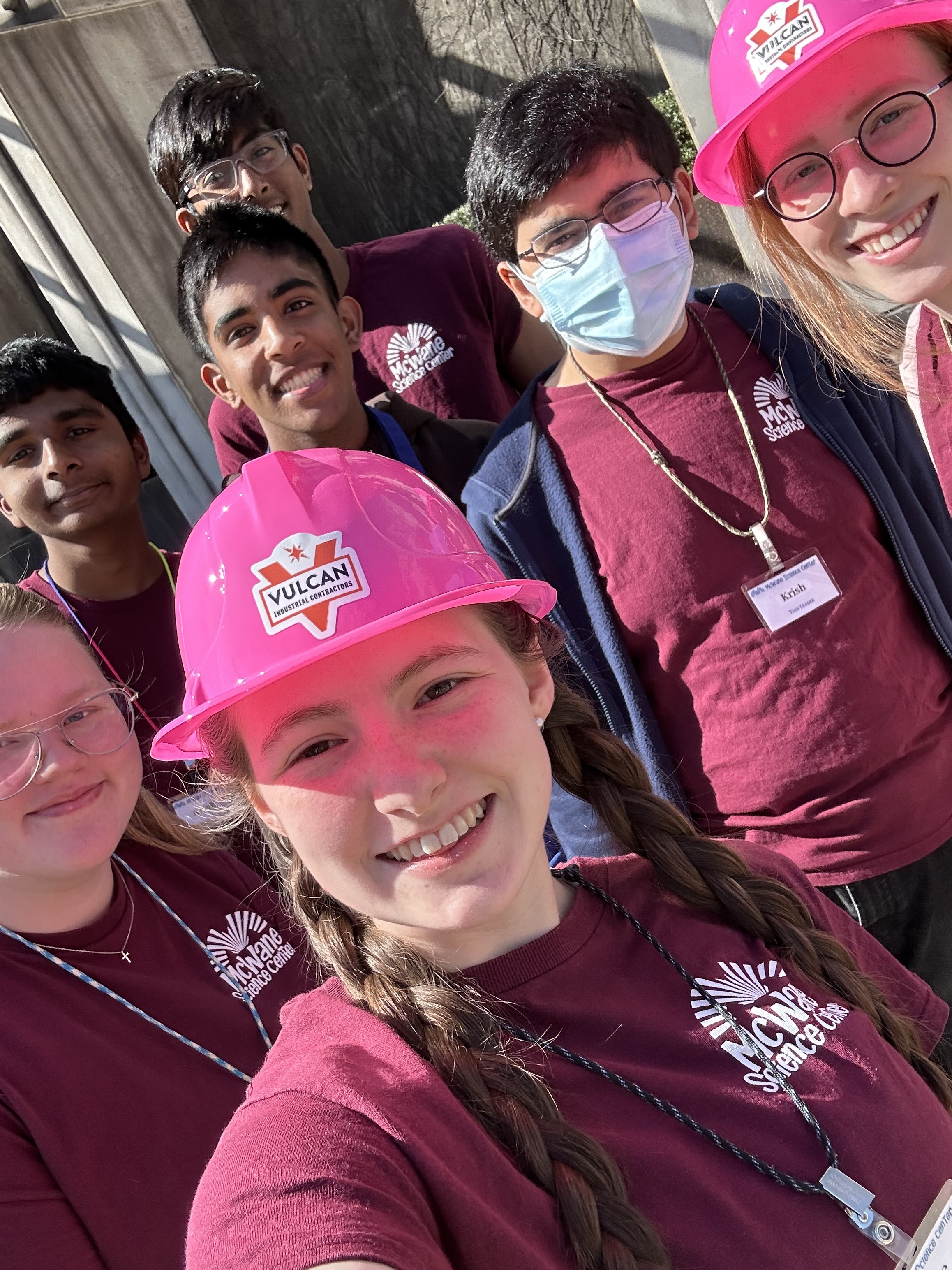 Teen volunteers at McWane Science Center in maroon shirts and pink hard hats smiling outside