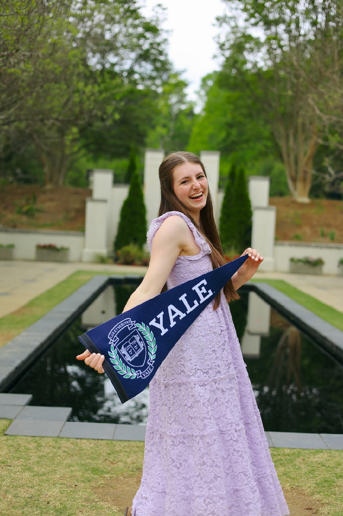 A woman in a lavender dress holds a Yale pennant by a reflecting pool at the children's science center garden.