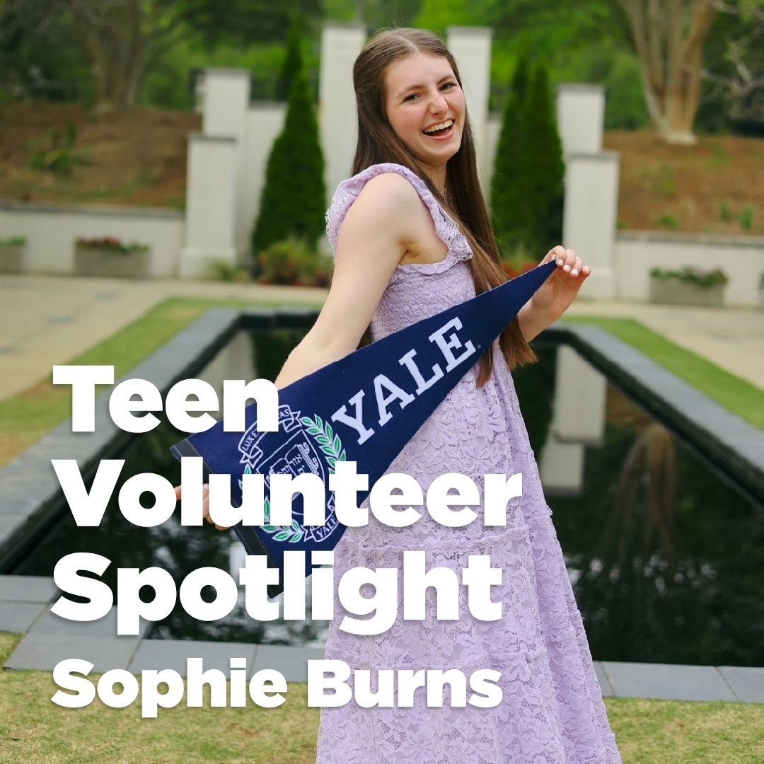 Teen volunteer at children's science center smiling outdoors, holding a Yale pennant by a reflecting pool.